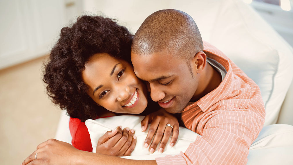 A married couple smiling and embracing on a couch, wedding rings visible
